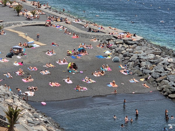 Playa de Radazul llena de bañistas disfrutando del sol sobre la arena negra protegida por escolleras.